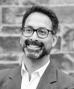 A black and white headshot of Dr. Andrew Pinto, who is smiling. He has short, dark hair, glasses, and a beard. 